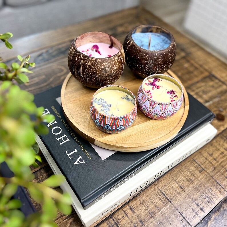 Peut inclure: Quatre bougies de diff&eacute;rentes couleurs et designs, dont une bougie bleue dans une coque de noix de coco, une bougie rose dans une coque de noix de coco, une bougie jaune dans une bo&icirc;te &agrave; motifs et une bougie jaune dans une bo&icirc;te &agrave; motifs. Les bougies sont dispos&eacute;es sur un plateau en bois avec un livre en dessous.