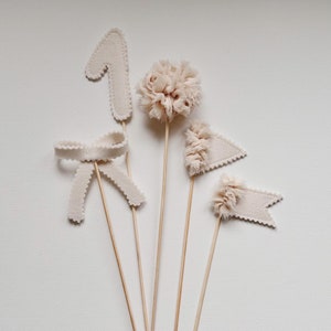 Puede incluir: Cinco decoraciones de fieltro beige en palos de madera. Las decoraciones incluyen un número uno, un lazo, un pompón y dos banderas.