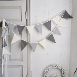 May include: A white and grey fabric bunting banner with triangle flags hanging on a white wall. The banner is decorated with a white wicker wreath and a white box with green plants.