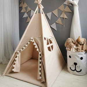 Puede incluir: Una tienda de campaña tipi de color beige con ribete de pompones blancos y una ventana. La tienda de campaña está instalada en una habitación con una alfombra blanca y una cesta de almacenamiento blanca con un diseño de cara de perro negro.