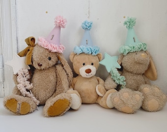 May include: Three plush toys wearing party hats with the number one on them. The toys are a brown bear, a brown bunny with a pink bow, and a brown bunny with a blue star.