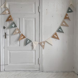 Puede incluir: Una guirnalda de banderines triangulares en colores neutros, deletreando un nombre. Los banderines son en tonos beige, verde y marrón, con letras blancas. La guirnalda cuelga frente a una puerta y una pared blancas.