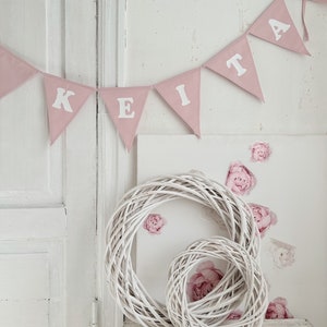 Puede incluir: Una pancarta de tela rosa con el nombre "KEITA" escrito en letras blancas. La pancarta está colgada en una pared blanca con una puerta blanca en el fondo. Hay dos coronas de mimbre blancas en un estante blanco debajo de la pancarta.