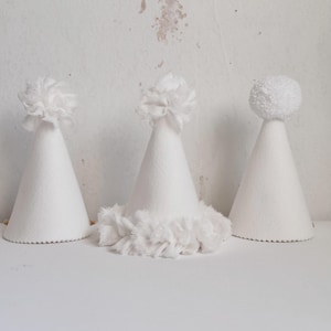 Pode incluir: Três chapéus de festa de feltro branco com diferentes enfeites. O primeiro chapéu tem um pompom branco, o segundo chapéu tem um babado branco e o terceiro chapéu tem uma flor branca.