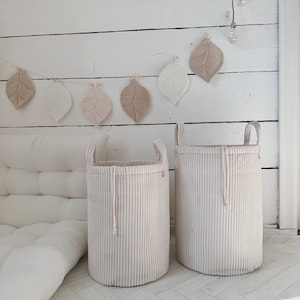 Op de afbeelding: Twee beige corduroy wasmanden met touwhandvatten en een trekkoord. De manden staan op een witte houten vloer met een witte muur op de achtergrond. Een guirlande van beige viltbladeren hangt aan de muur.