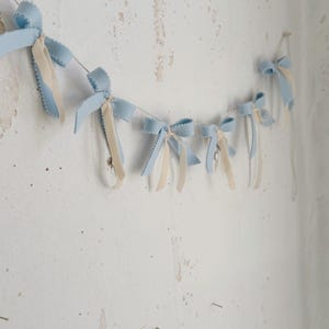 May include: A garland of light blue felt bows with white satin ribbon tails hanging on a white wall.