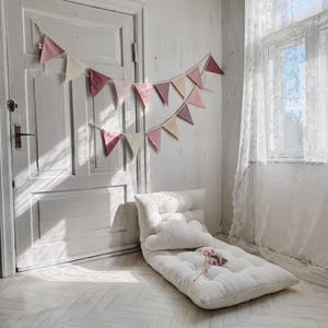 May include: A white, fluffy floor cushion with a white pillow and a pink decorative pillow. The cushion is in a room with white walls and a white door. A pink and white bunting hangs above the cushion.