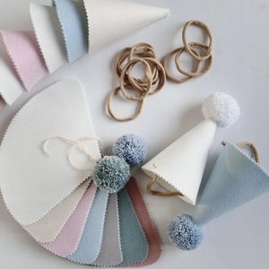 Pode incluir: Um conjunto de chapéus de festa de feltro em várias cores, incluindo branco, azul claro e rosa. Os chapéus são em forma de cone e têm um pompom no topo. Há também várias amostras de tecido de feltro em diferentes cores, incluindo branco, rosa claro, azul claro, cinza e marrom. As amostras de tecido estão dispostas em forma de leque. Há também várias elásticos de cabelo marrons.