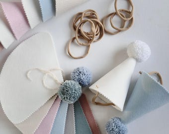 Pode incluir: Um conjunto de chapéus de festa de feltro em várias cores, incluindo branco, azul claro e rosa. Os chapéus são em forma de cone e têm um pompom no topo. Há também várias amostras de tecido de feltro em diferentes cores, incluindo branco, rosa claro, azul claro, cinza e marrom. As amostras de tecido estão dispostas em forma de leque. Há também várias elásticos de cabelo marrons.