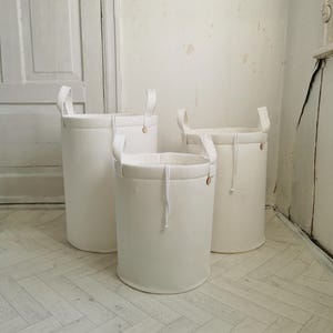 Op de afbeelding: Drie witte opbergmanden van stof met handvatten en een touwdetail. De manden staan in een rij op een lichte houten vloer.