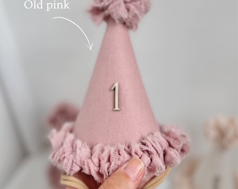 May include: A conical party hat in a dusty old pink shade, featuring a frayed fabric ruffle at the base and a matching pom-pom at the top. The front of the hat displays the number 1 in a light-colored material. A thin elastic band is attached.