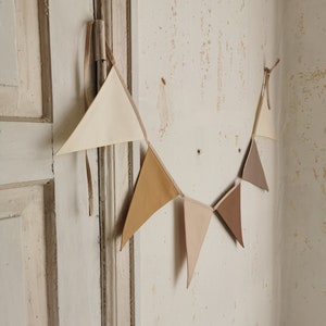 May include: A string of fabric pennants in beige, brown, and light brown, hanging on a white wall. The pennants are triangular in shape and are connected by string.
