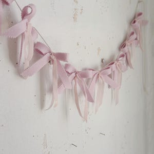 May include: A pink felt ribbon garland with bows hanging on a white wall.