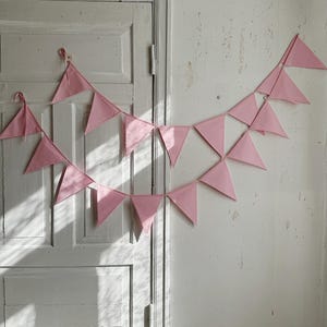 Può includere: Una ghirlanda di bandierine rosa in tessuto è appesa a una porta e a un muro bianchi. Le bandiere triangolari sono distanziate in modo uniforme, creando una decorazione festosa. La porta e il muro hanno un aspetto invecchiato e vintage.