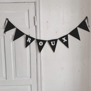 May include: Black fabric banner with white letters spelling out the word "ROUX" hanging on a white wall.