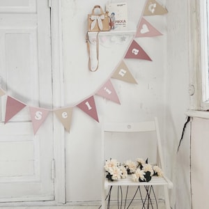Puede incluir: Un banderín de tela rosa y beige con la palabra "Selina" escrita en letras blancas. El banderín está colgado en una pared blanca con una puerta blanca y una silla blanca con un ramo de flores blancas.