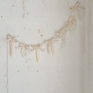 May include: A garland of cream-colored bows hangs against a textured white wall. The bows are evenly spaced and connected by a delicate string, creating a simple, elegant decoration. The wall has visible imperfections.