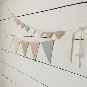 May include: Two pastel-colored bunting banners hanging against a white, weathered wood wall. The pennants are in shades of pink, gray, and cream, strung on natural-colored twine, creating a soft, decorative display.