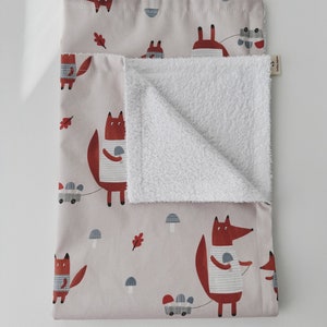 May include: A soft, pink and white baby blanket with a fox print. The fox is wearing a striped shirt and carrying a mushroom. The blanket has a white terry cloth side and a pink cotton side with a fox print.
