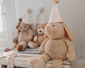 May include: Three plush toys wearing party hats. A brown bunny rabbit with long ears sits on a white wooden crate. A brown teddy bear and a brown bunny rabbit sit behind the bunny rabbit.