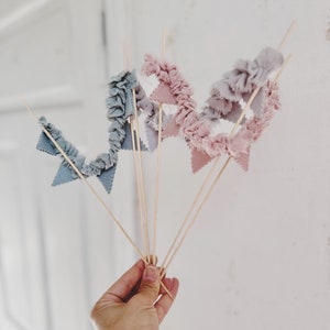 Puede incluir: Un conjunto de cinco banderas de tela en palos de madera. Las banderas son de dos colores: azul claro y rosa claro. Las banderas están fruncidas y tienen forma triangular.