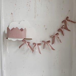 May include: A pink felt crown with the name "Linda" embroidered on it, hanging on a white cloud-shaped shelf. A pink felt garland with bows hangs above the crown.