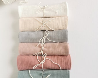 May include: A stack of seven rolled, soft-looking fabric cloths in various pastel colors, including white, beige, gray, pink, and teal. Each cloth is tied with a white string. A spool of white string sits at the top left.