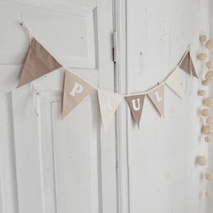 Op de afbeelding: Een witte en bruine stoffen banner met de naam "PAUL" in witte letters. De banner hangt aan een witte muur.