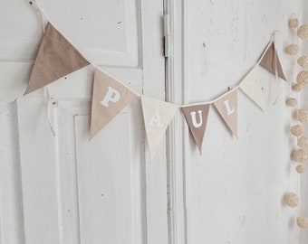 May include: A white and brown fabric banner with the name "PAUL" spelled out in white letters. The banner is hanging on a white wall.