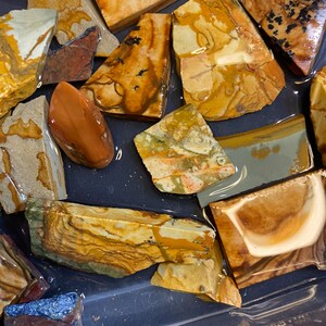 Puede incluir: Una colecci&oacute;n de piedras de &aacute;gata pulidas multicolores con patrones marrones, amarillos, blancos y naranjas. Las piedras est&aacute;n dispuestas sobre una superficie gris oscura.