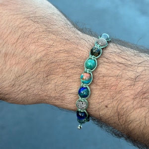 May include: A green and blue beaded bracelet with silver accents. The bracelet is made with different types of stones and is tied with a green cord.