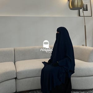 May include: A person wearing a navy blue niqab and a black dress, seated on a light-colored sofa. The niqab covers the face, with only the eyes visible. The image includes the logo "niqabi style".