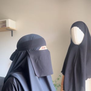 Puede incluir: Dos niqabs negros, uno con velo facial y otro sin. El niqab con el velo facial tiene una banda negra alrededor de la frente.