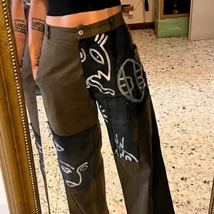 Può includere: Un paio di pantaloni cargo verde scuro con disegni astratti in bianco e nero e una grafica di un viso sulla gamba sinistra.