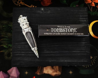 May include: A silver lip gloss tube with a crown-shaped top and a black label that reads "Petals & Plumes Tombstone Hydrating Lip Gloss | Sweet Coconut | 0.16oz/4.4mL"