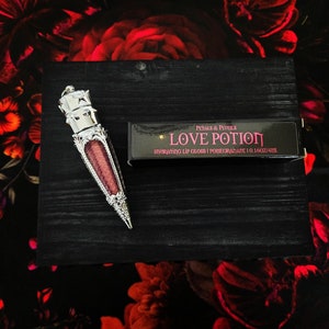 Puede incluir: Un tubo de brillo de labios plateado y negro con una tapa decorativa en forma de corona. El tubo está etiquetado "LOVE POTION HYDRATING LIP GLOSS | POMEGRANATE 0.16OZ/4.4ML" en texto rosa y blanco.