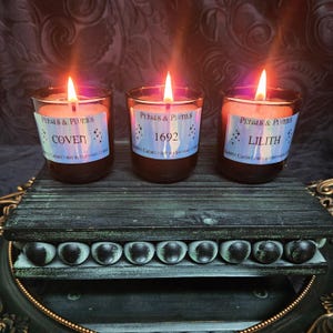 Puede incluir: Tres tarros de velas de vidrio marrón con etiquetas negras. Las etiquetas tienen un diseño holográfico y dicen "Petals & Plumes" y los nombres de las fragancias: "Covet", "1692" y "Lilith". Las velas están sobre una plataforma de madera oscura con un borde decorativo.