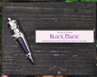 May include: A cone-shaped lip mud container with a metallic silver cap and a deep purple textured body, positioned next to a rectangular box labeled Petals & Plumes Black Magic Shimmer Matte Lip Mud Chocolate Scent 0.16oz/4ml.