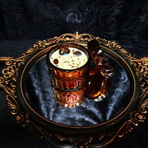 May include: A brown glass candle jar with a gold lid, filled with a white candle and topped with decorative brown and gold accents. The candle is sitting on a gold and black decorative tray.