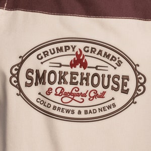 May include: A tan apron with a brown embroidered logo that says "Grumpy Gramps Smokehouse & Backyard Grill, Cold Brews & Bad News".