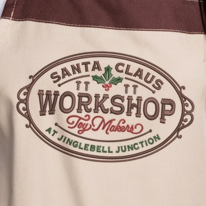 May include: A beige apron with a brown border features a vintage-style logo that reads "Santa Claus Workshop at Jinglebell Junction Toy Makers".
