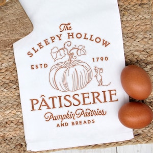 May include: White kitchen towel with a brown embroidered design of a pumpkin, a mouse, and the text "The Sleepy Hollow Pâtisserie Pumpkin Pastries and Breads EstD 1790".