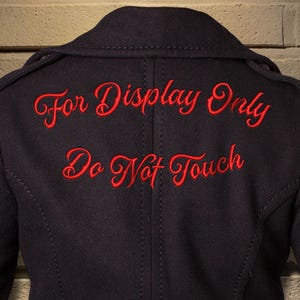 Puede incluir: Chaqueta negra con texto bordado en rojo "For Display Only" y "Do Not Touch". La chaqueta tiene un diseño cruzado y un corte entallado. El texto está en cursiva.