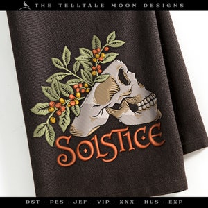 May include: A black kitchen towel with an embroidered design of a skull with a floral wreath and the word "Solstice" in orange.