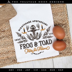 May include: White kitchen towel with a black and gold embroidered design. The design features a frog skeleton with candles, rosemary sprigs, and the text "Nightshade Apothecary 1692 171 Frog & Toad Parts for Potions".