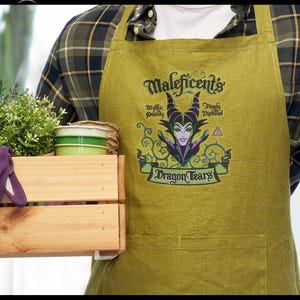 May include: A green apron featuring the text "Maleficent's Dragon Tears" with an illustration of Maleficent. The apron has a pocket and is worn over a plaid shirt. A wooden crate with plants and small green and white containers is in the foreground.