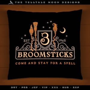 Pode incluir: Uma almofada preta com um design bordado em dourado que apresenta três vassouras, uma lua crescente e o texto "EST 1046 BROOMSTICKS COME AND STAY FOR A SPELL".