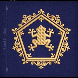 Może przedstawiać: Złoty haft przedstawiający żabę w sześciokątnej ramie z ozdobnymi detalami. Projekt ma 12 cm wysokości.