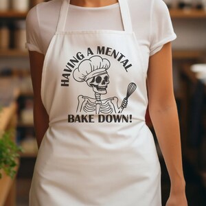 Op de afbeelding: Witte schort met de tekst "HAVING A MENTAL BAKE DOWN!" in zwart. Het ontwerp toont een skelet met een koksmuts en een garde. De schort is gemaakt van een gladde stof, waarschijnlijk katoen of polyester.