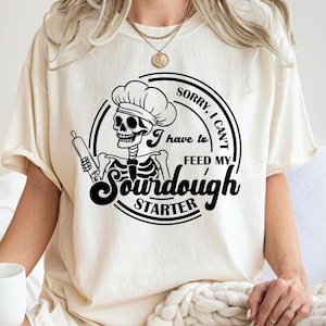 Może przedstawiać: Kremowa koszulka z czarną grafiką przedstawiającą szkielet kucharza trzymającego wałek do ciasta z napisem "Sorry, I can't feed my sourdough starter". Projekt jest zamknięty w okrągłej ramie.